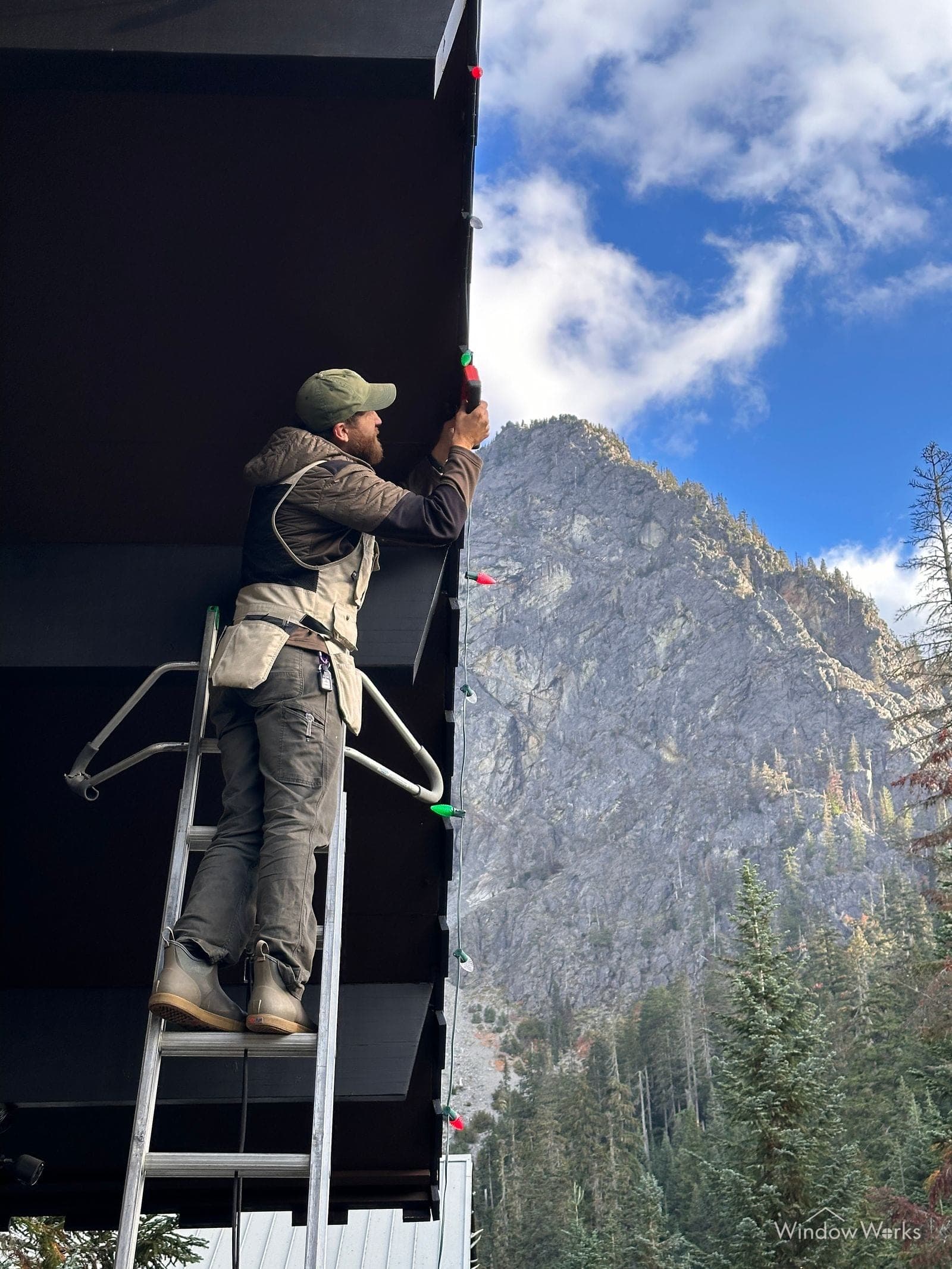 A technician on a ladder hanging lights at an eave with a mountain face behind.