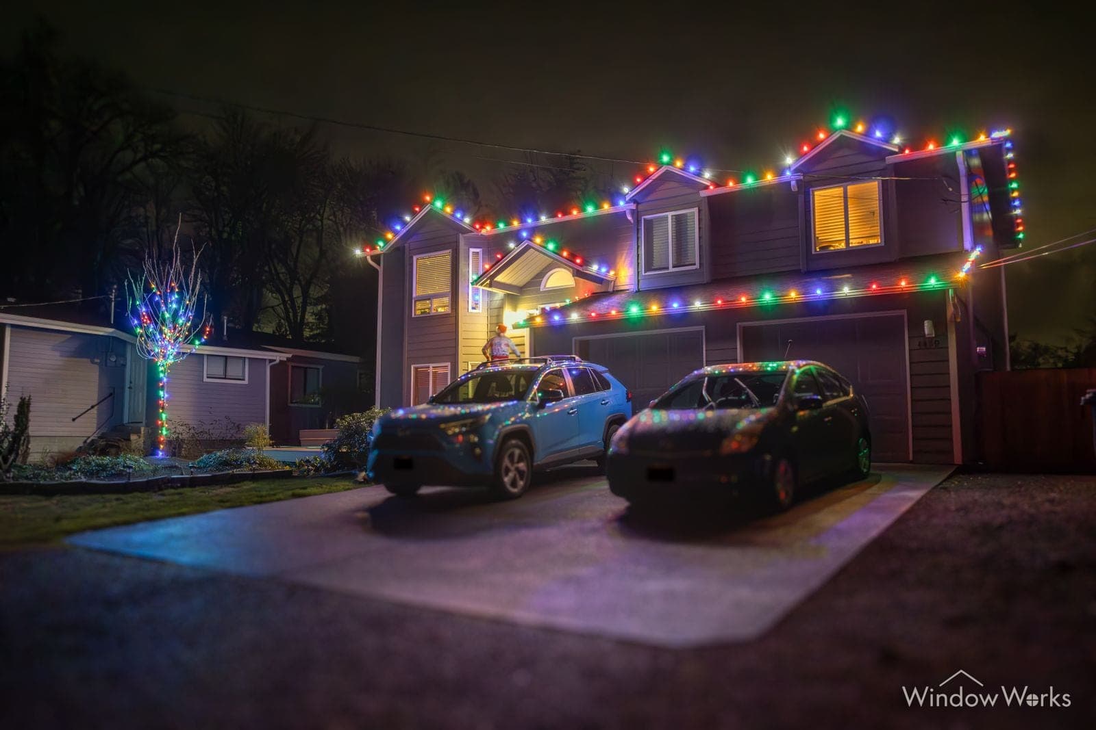A two-story home in multicolor bulbs with two cars on the drive and a wrapped tree beside it.