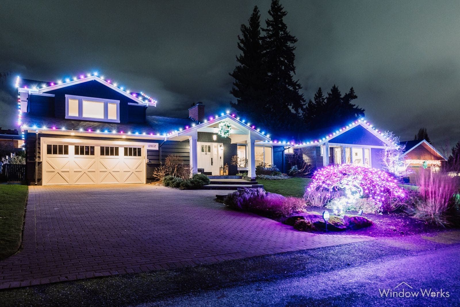 A single-story home in warm white bulbs with a magenta uplit shrub and color washing across the wet drive.