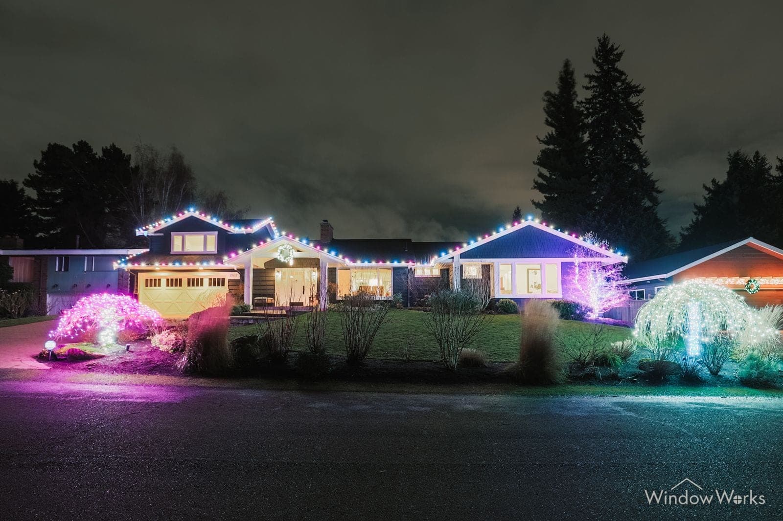 A rambler at night in warm white with pink and teal uplit shrubs across the front.