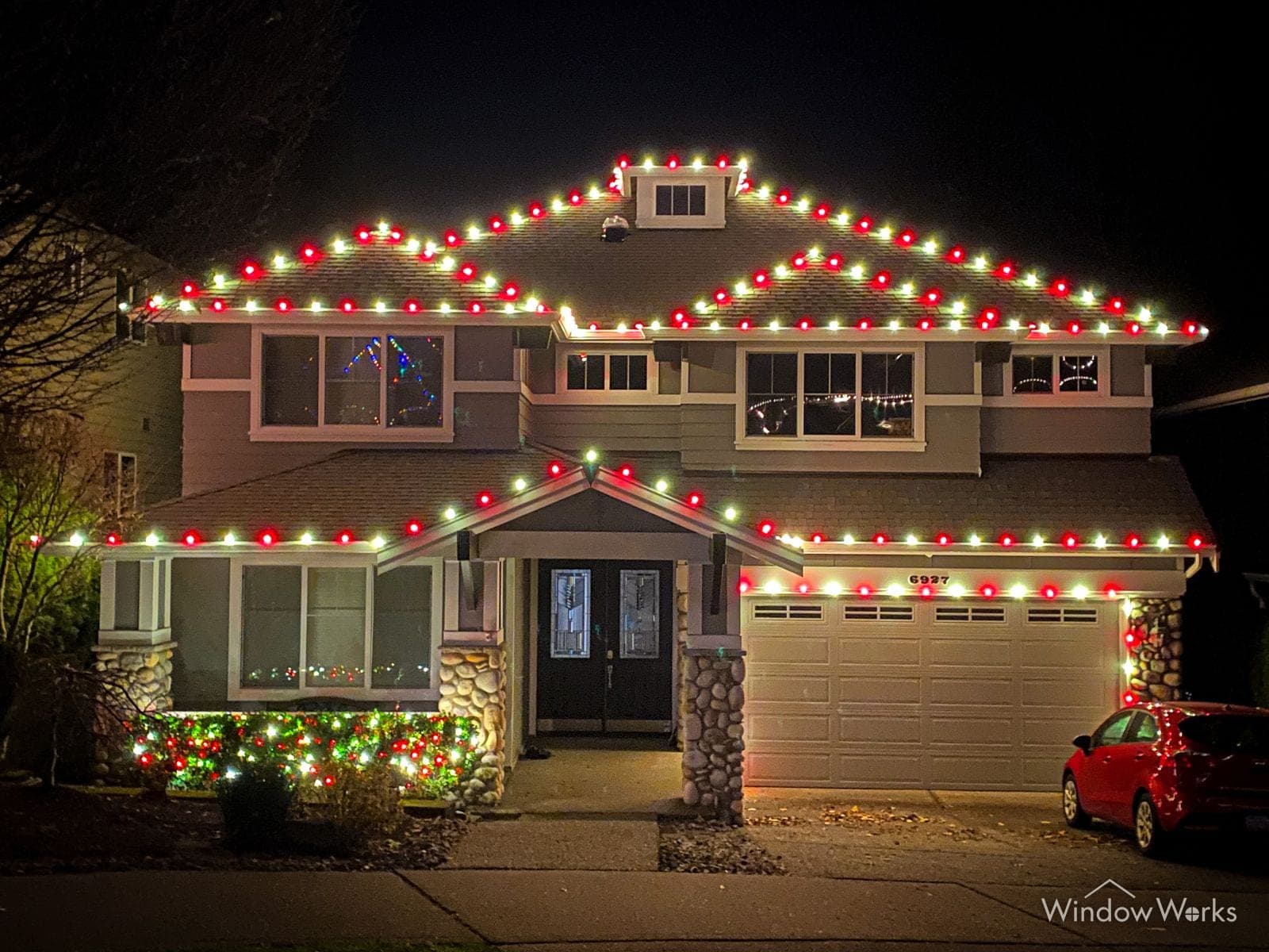 A two-story home in red and green bulbs with lit beds and a red car on the drive.
