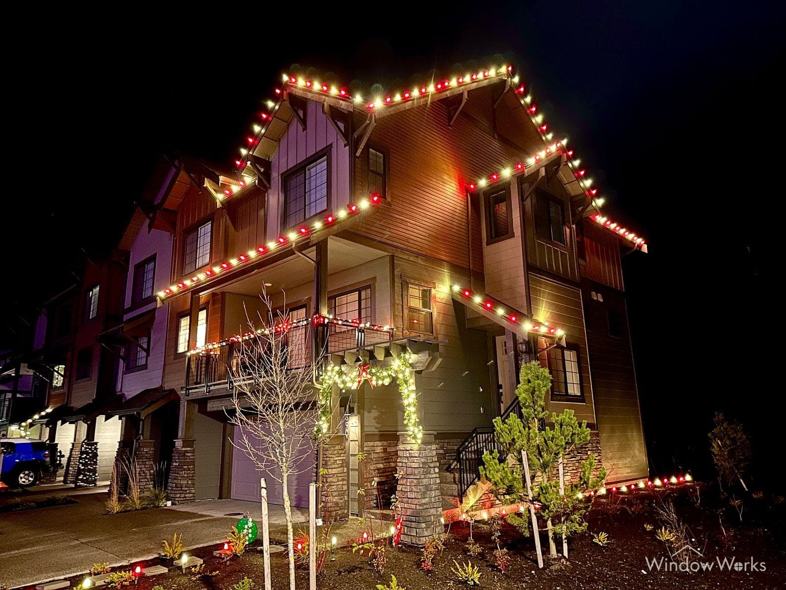 A tall three-story home in red, green and warm white bulbs, porch rails and beds lit below.