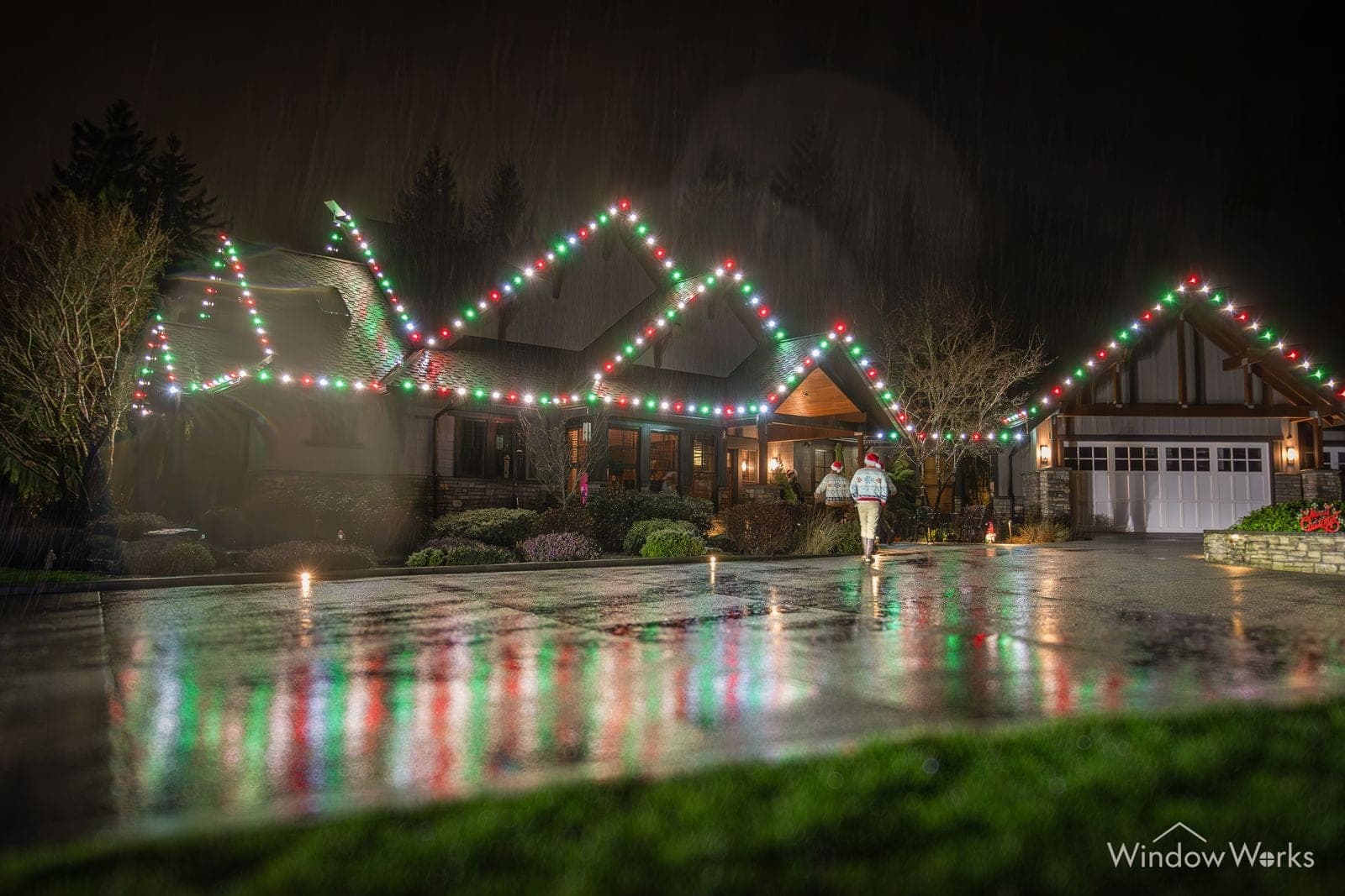 A steep-gabled home in red, green and warm white bulbs, every peak reflected in the wet street.