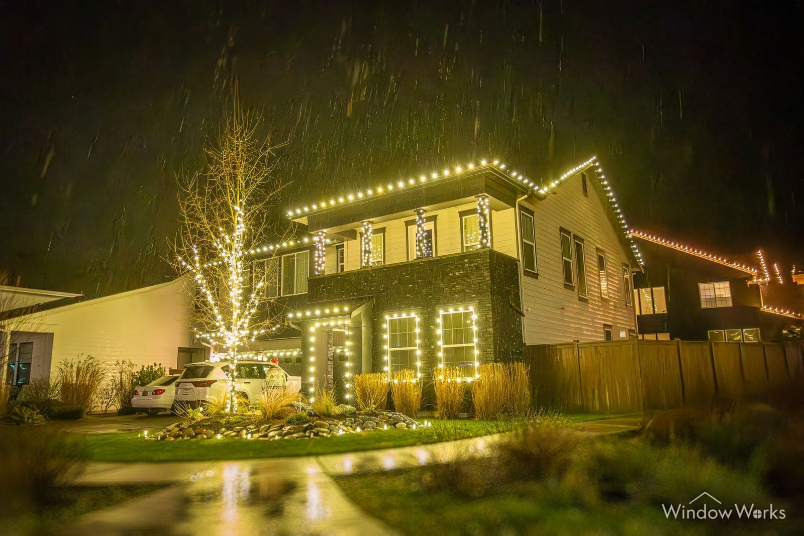 A two-story home in warm white bulbs with a white-lit birch by the drive and lit garage trim.