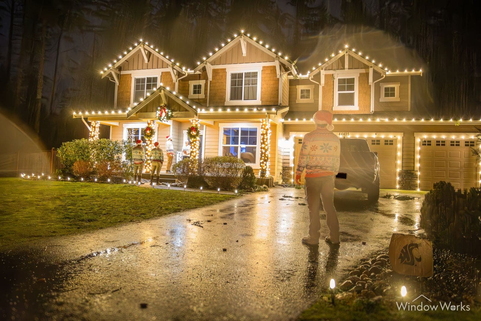 A home in warm white bulbs with wreaths in the windows and the owner crossing the wet drive.