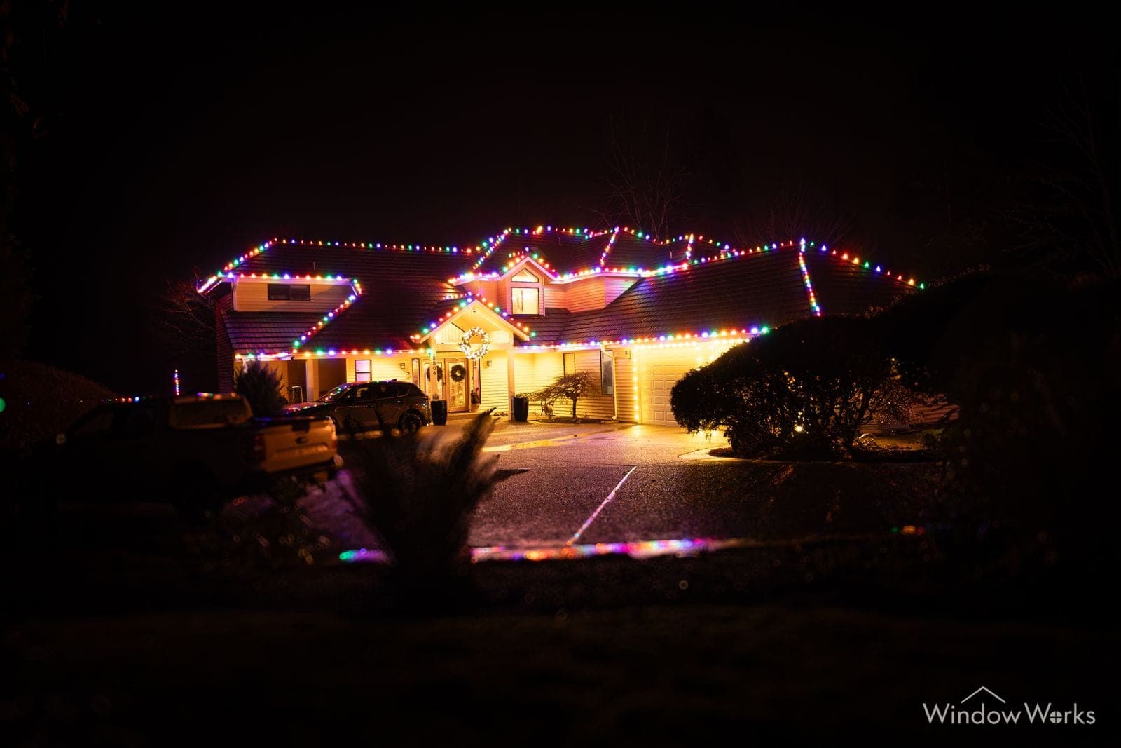 A low home across a broad lawn at night, roofline in multicolor bulbs with a lit tree at the door.