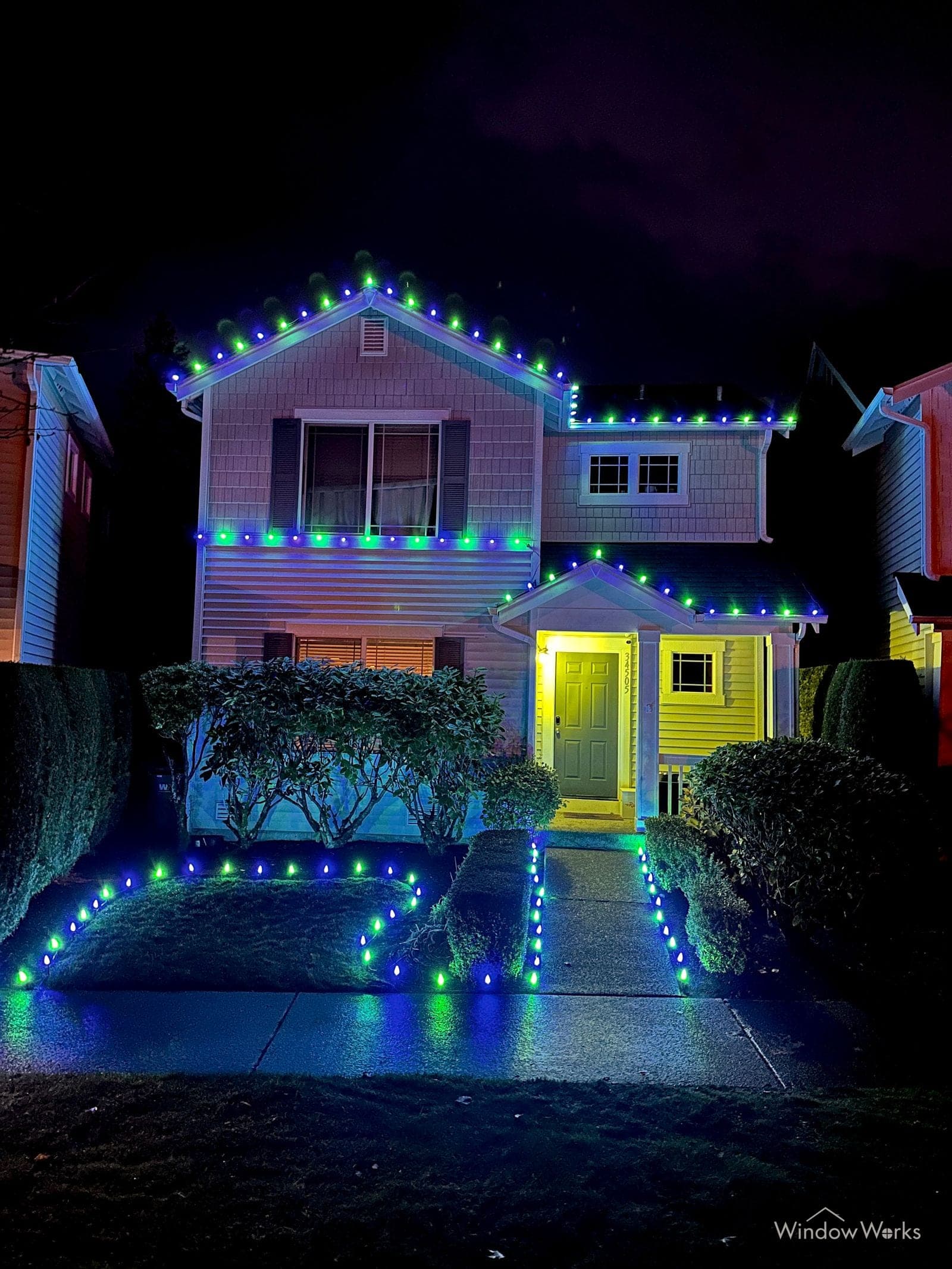 A home lit blue and green along the rooflines with a lit path running between hedges to the front door.