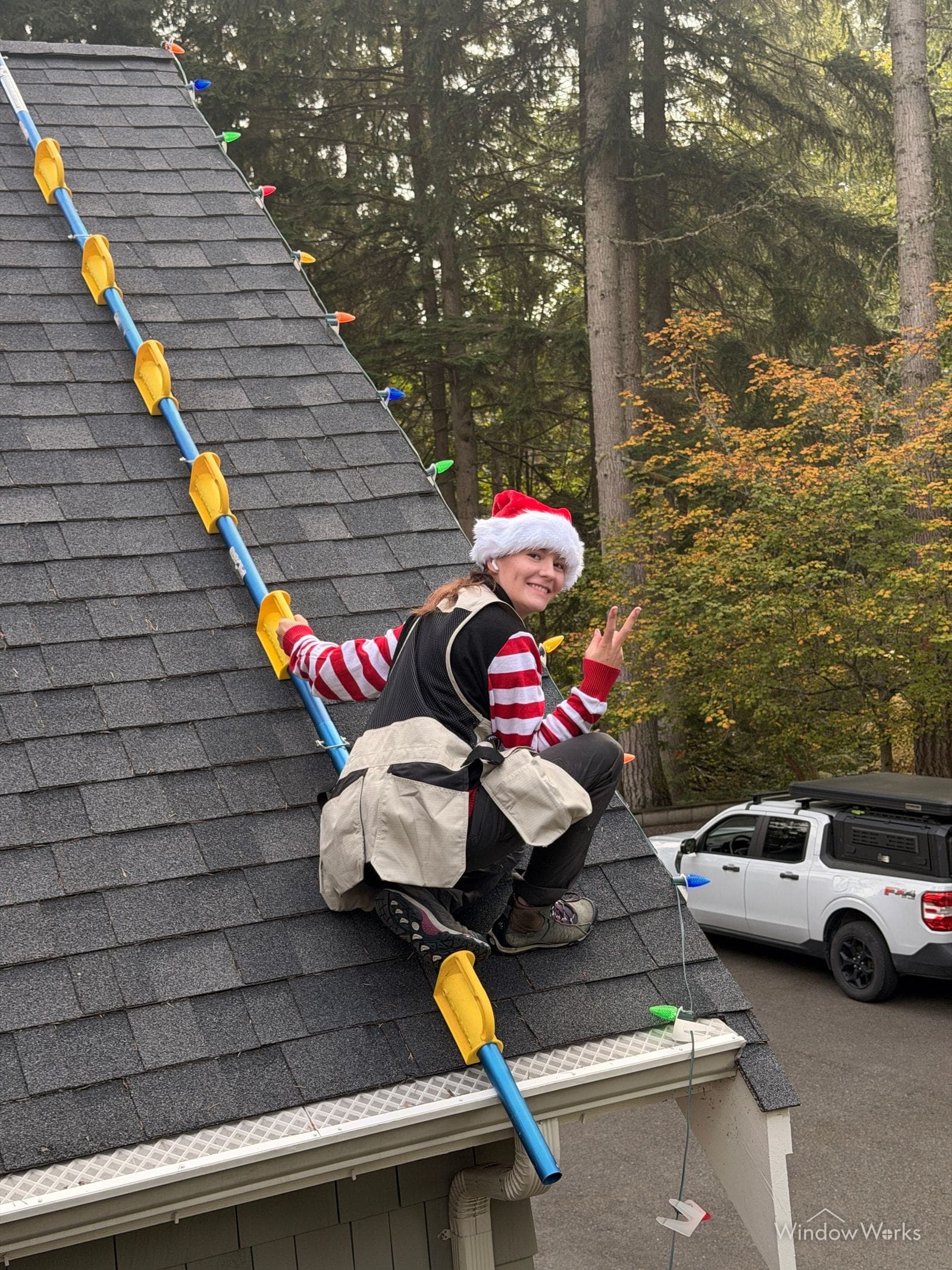A WindowWorks technician in a Santa hat and harness clipping holiday lights along a roofline, waving from the roof