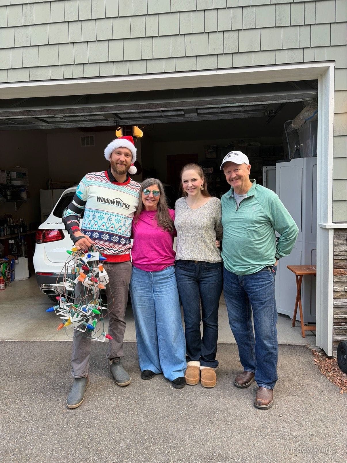 A WindowWorks technician in a company holiday sweater and Santa hat holding strands of holiday lights beside three smiling homeowners in their garage