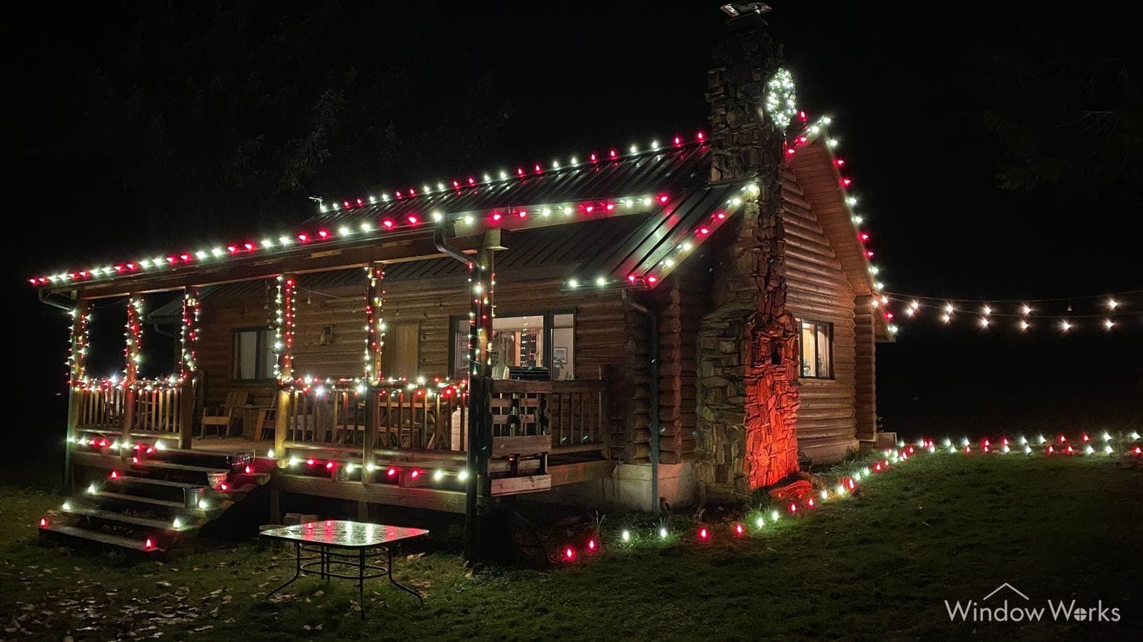 A log home at night with red, green and warm white bulbs along the porch roof and out along the fence line.