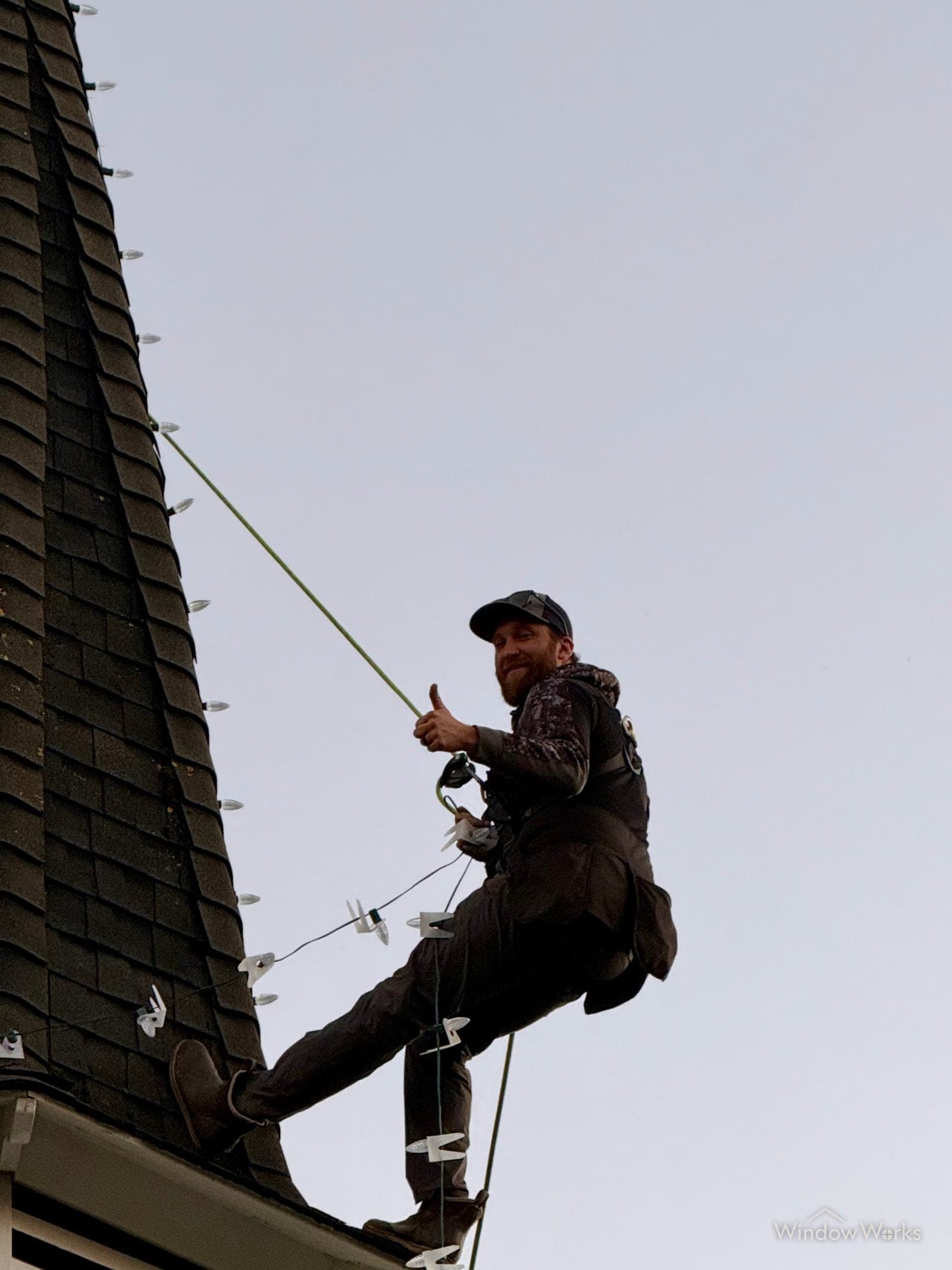 A WindowWorks technician rappelling down a steep church steeple on rope, giving a thumbs up beside the lights they are installing