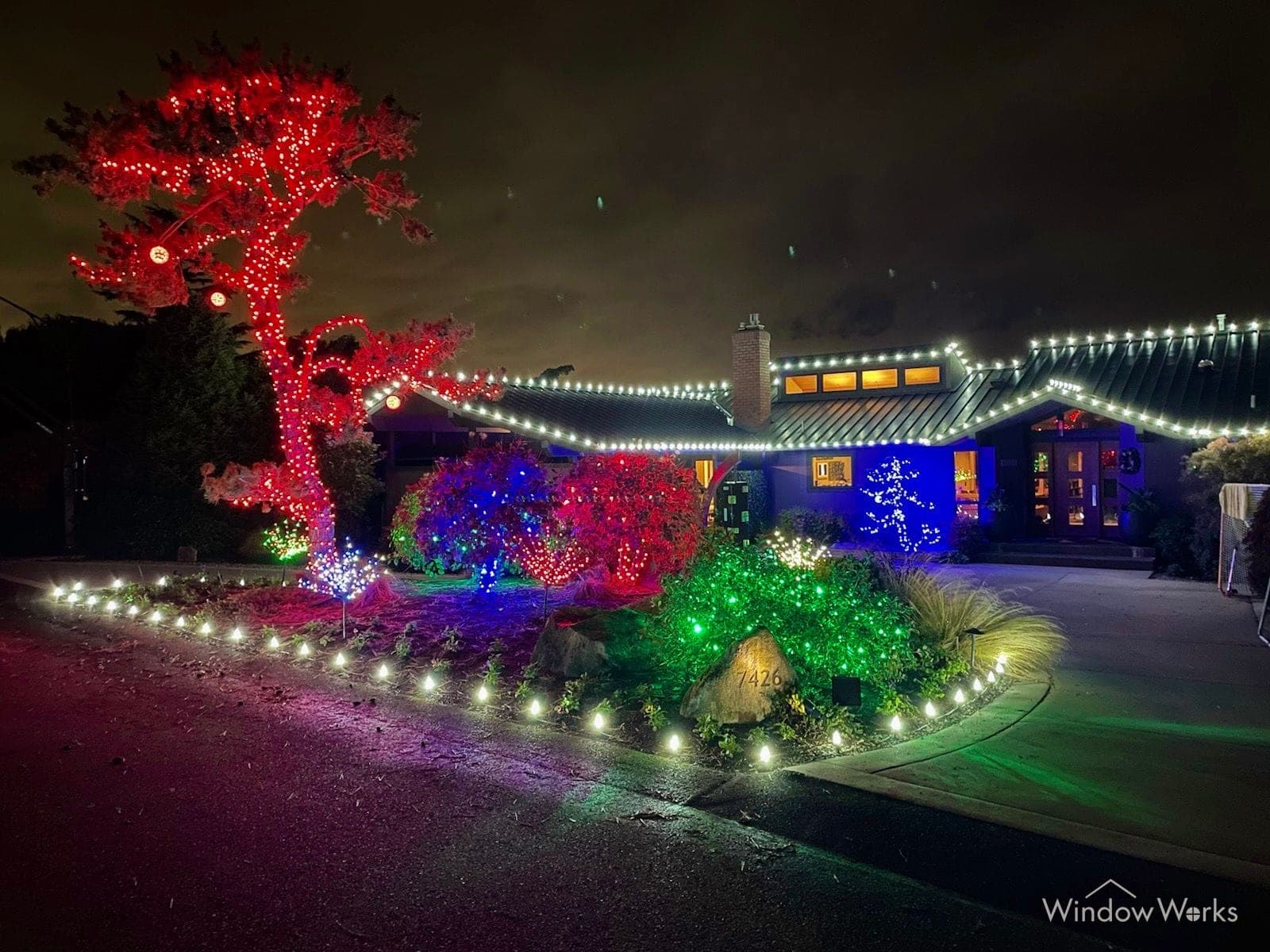 A low home lit warm white with a red-wrapped tree above the drive and colored lit shrubs running the length of the front beds.
