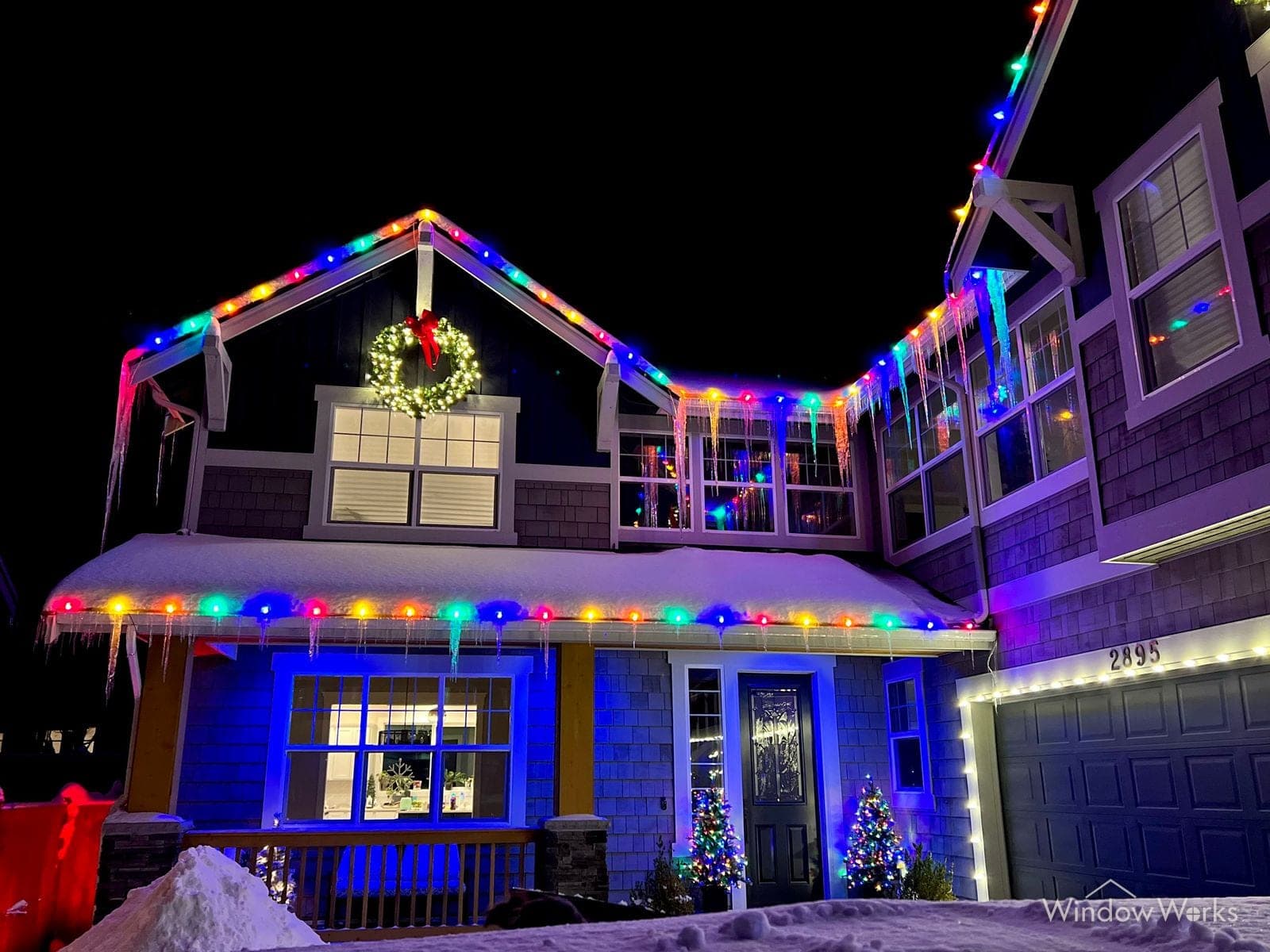 A snow-banked home lit in multicolor C9 bulbs with blue uplighting on the walls and lit trees either side of the front door.