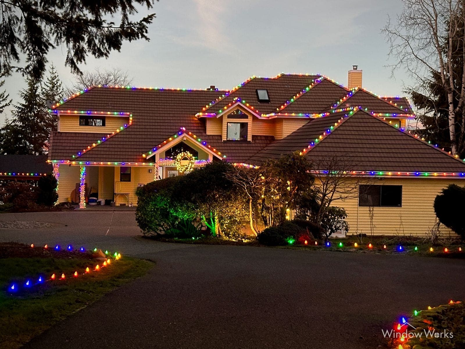 A tile-roofed home at dusk with multicolor bulbs following every roof edge and dormer, and a lit path running out to the drive.
