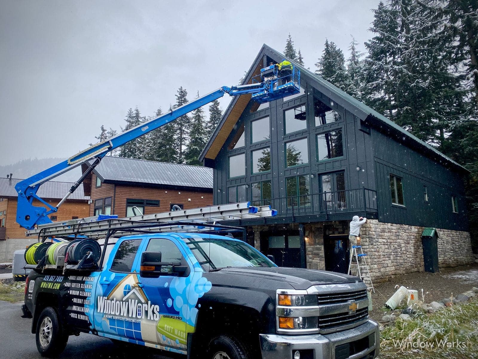 The WindowWorks truck and a boom lift working the gable of a modern timber lodge, firs behind.