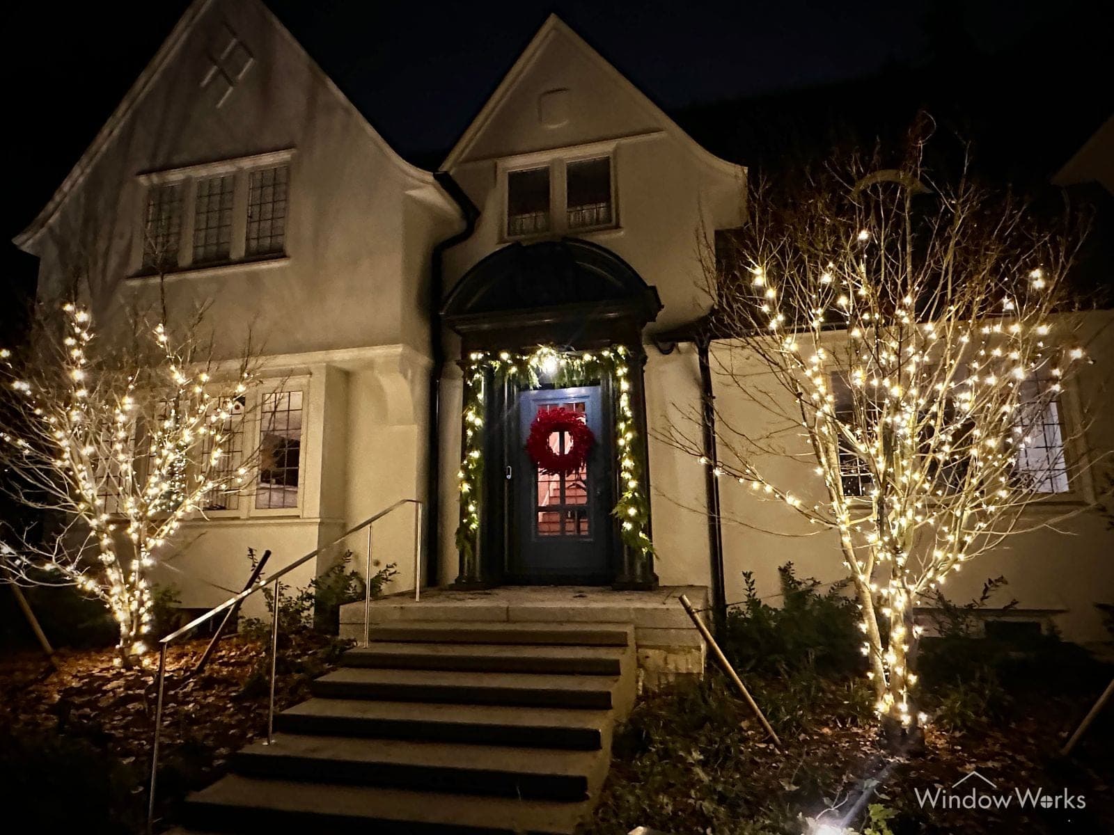 A tudor home at night with two bare trees wrapped in warm white either side of the steps and a lit wreath on the front door.