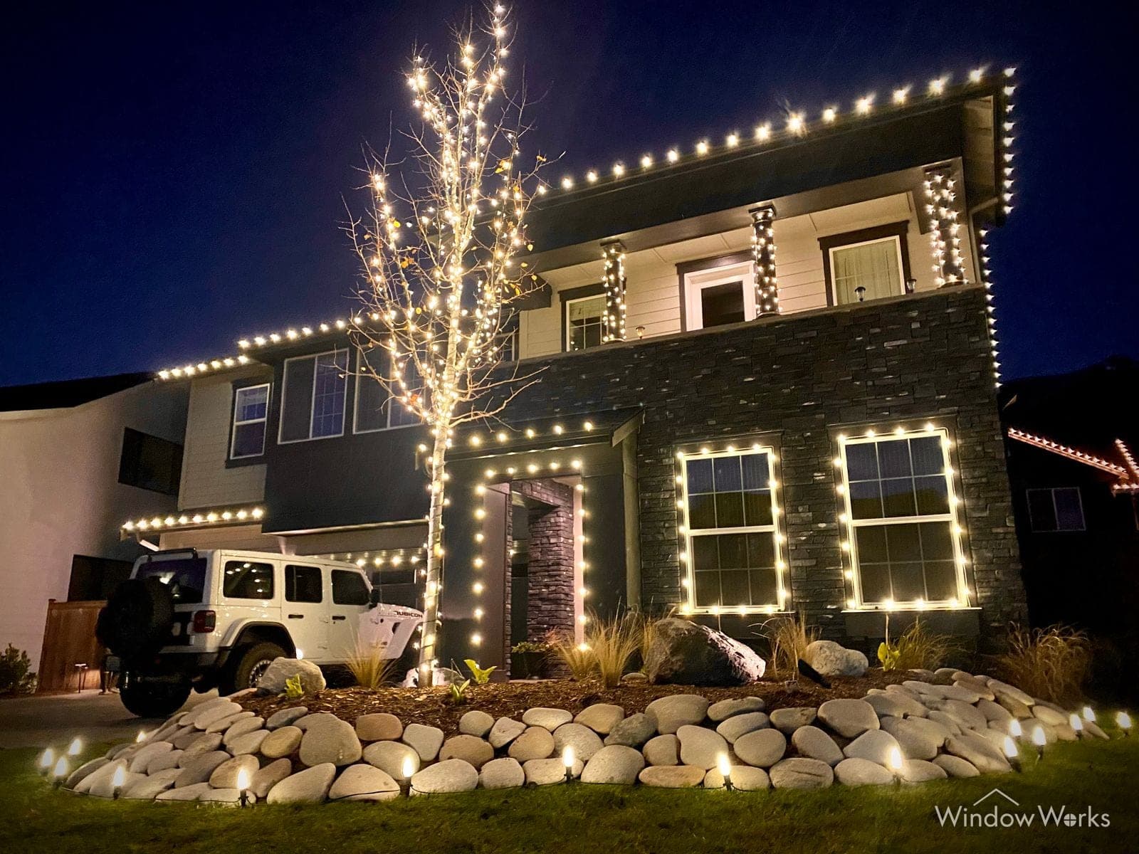 A two-story home outlined in warm white bulbs with a bare tree wrapped from trunk to branch tips, boulders and a lit bed along the drive.