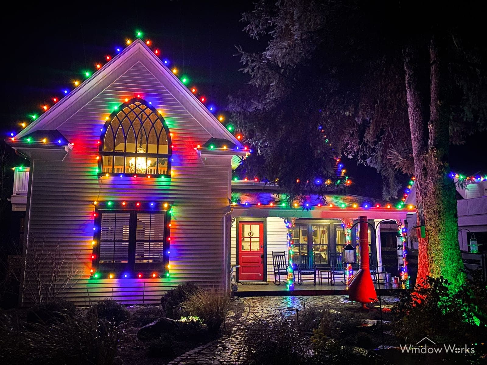 A white gabled home at night outlined in multicolor bulbs around every window, gable and porch, the arched front window glowing.