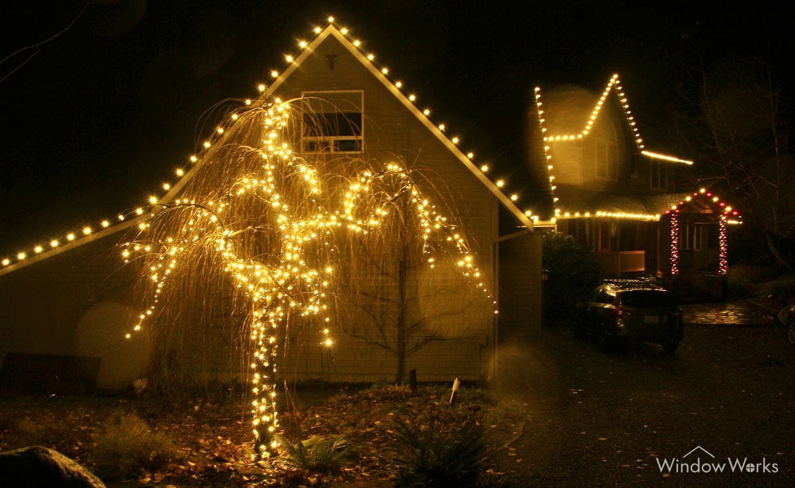 A weeping tree lit in warm white beside a home whose gable and eave carry the same warm bulbs.