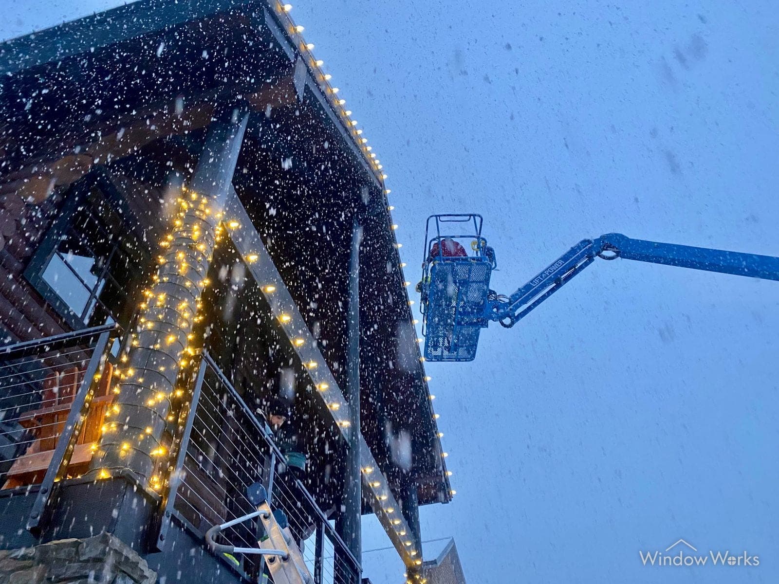 A WindowWorks technician in a boom lift installing holiday lights along a high gable in falling snow.