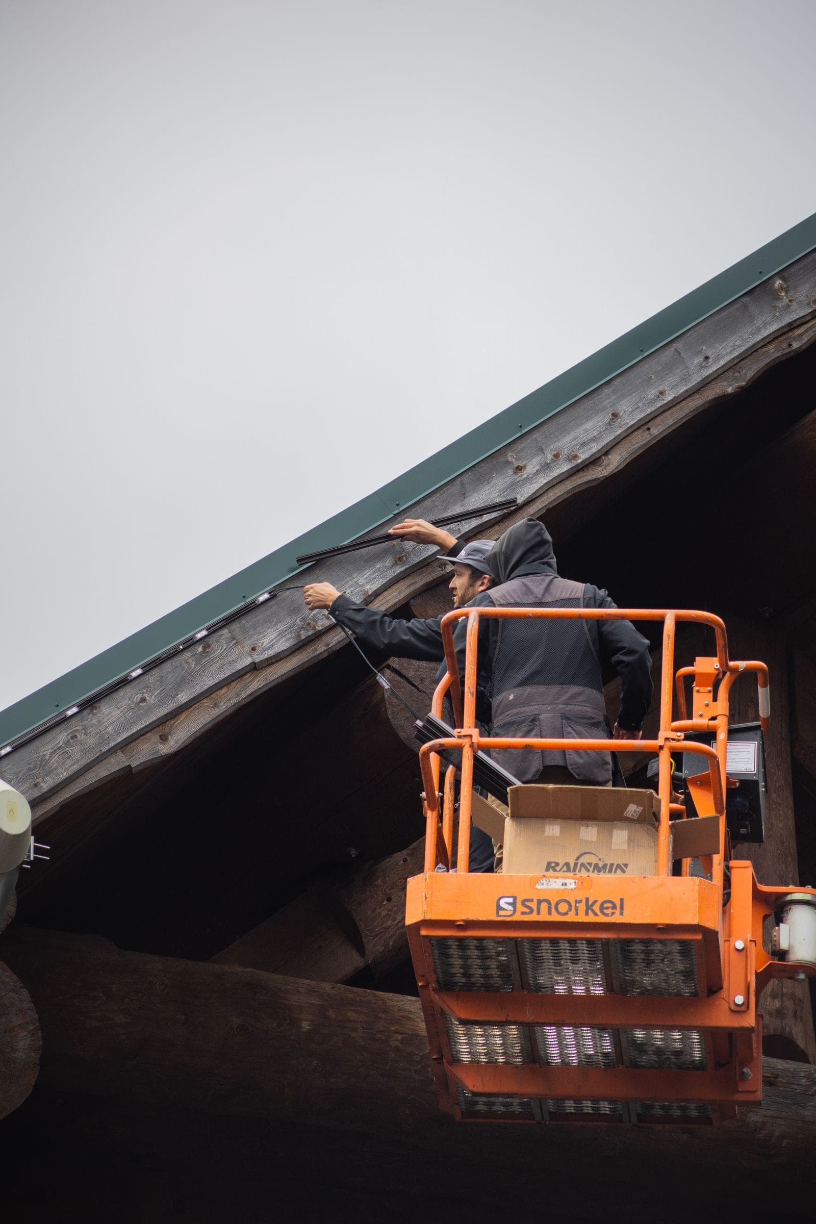 A technician in a boom lift running a permanent lighting line along the timber gable of a lodge on an overcast day.
