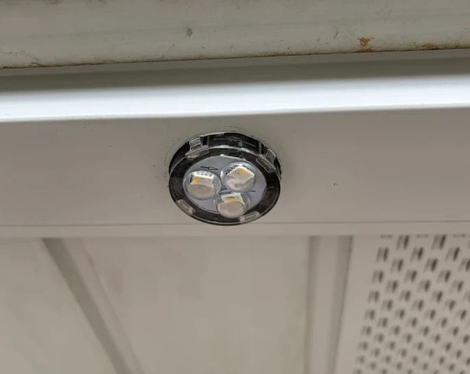 A close view of a permanent puck light seated flush into a white soffit, its three diodes visible behind the lens.