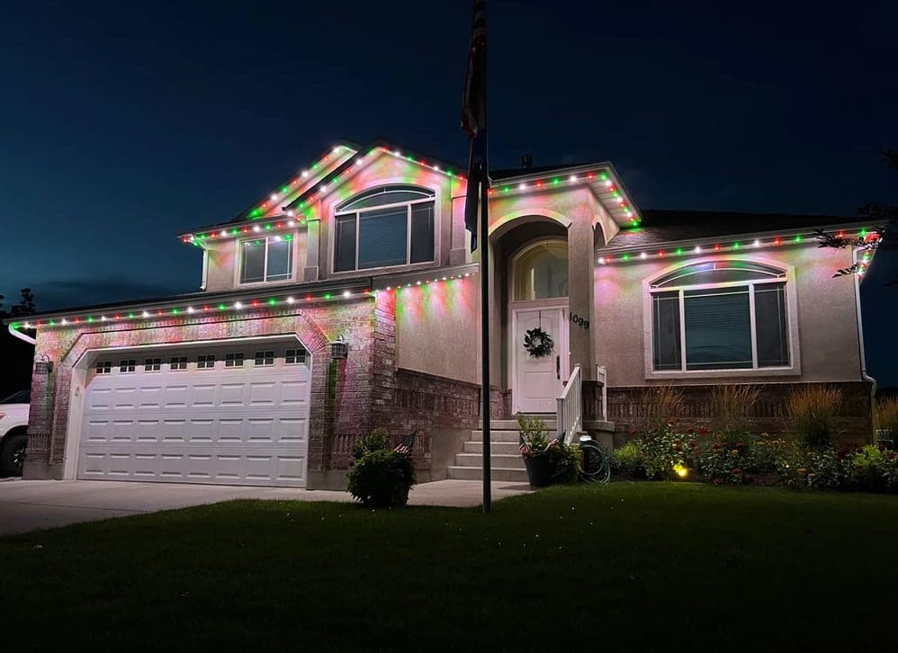 A stucco two-story home at dusk with permanent lighting running red, green and warm white along every roofline and dormer.