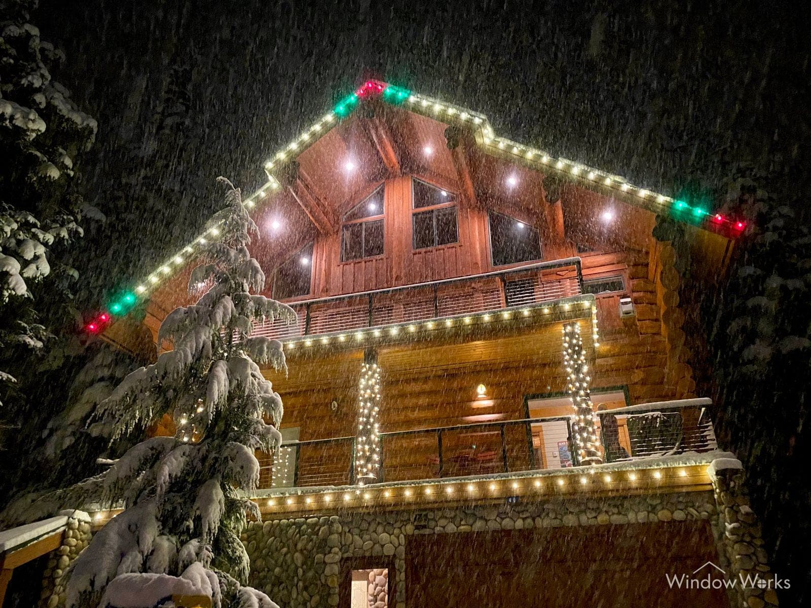 A mountain chalet in falling snow at night, rooflines and balcony lit with multicolor and warm white holiday lights, snow-loaded evergreens beside it.