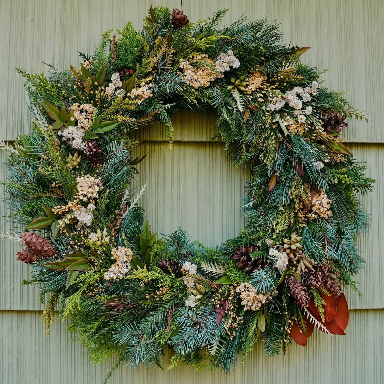 A classic evergreen wreath, full and round, dressed with berries, cones and dried flowers on green siding.