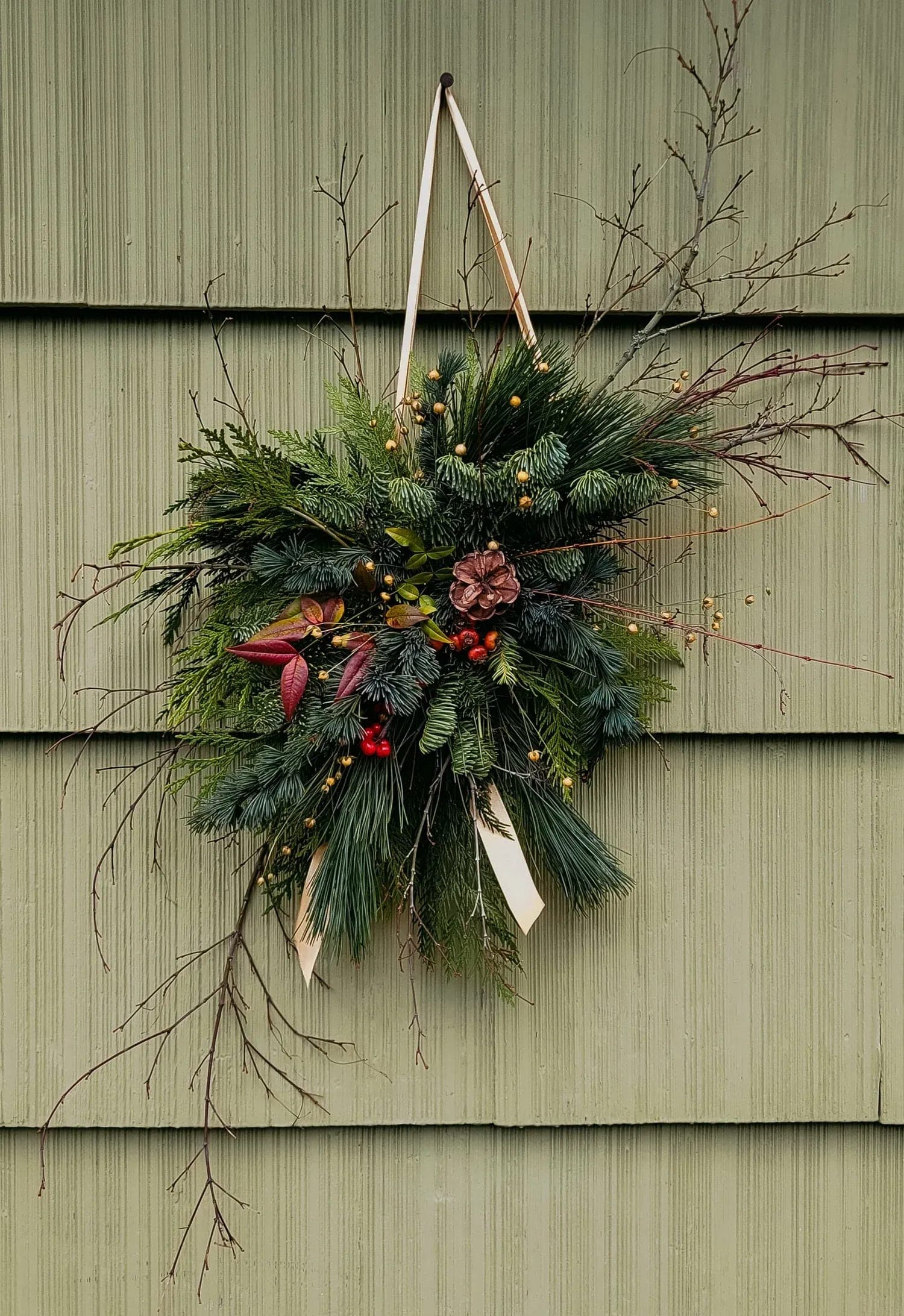 A door swag of layered evergreen and cedar with a trailing ribbon, hung on green siding.