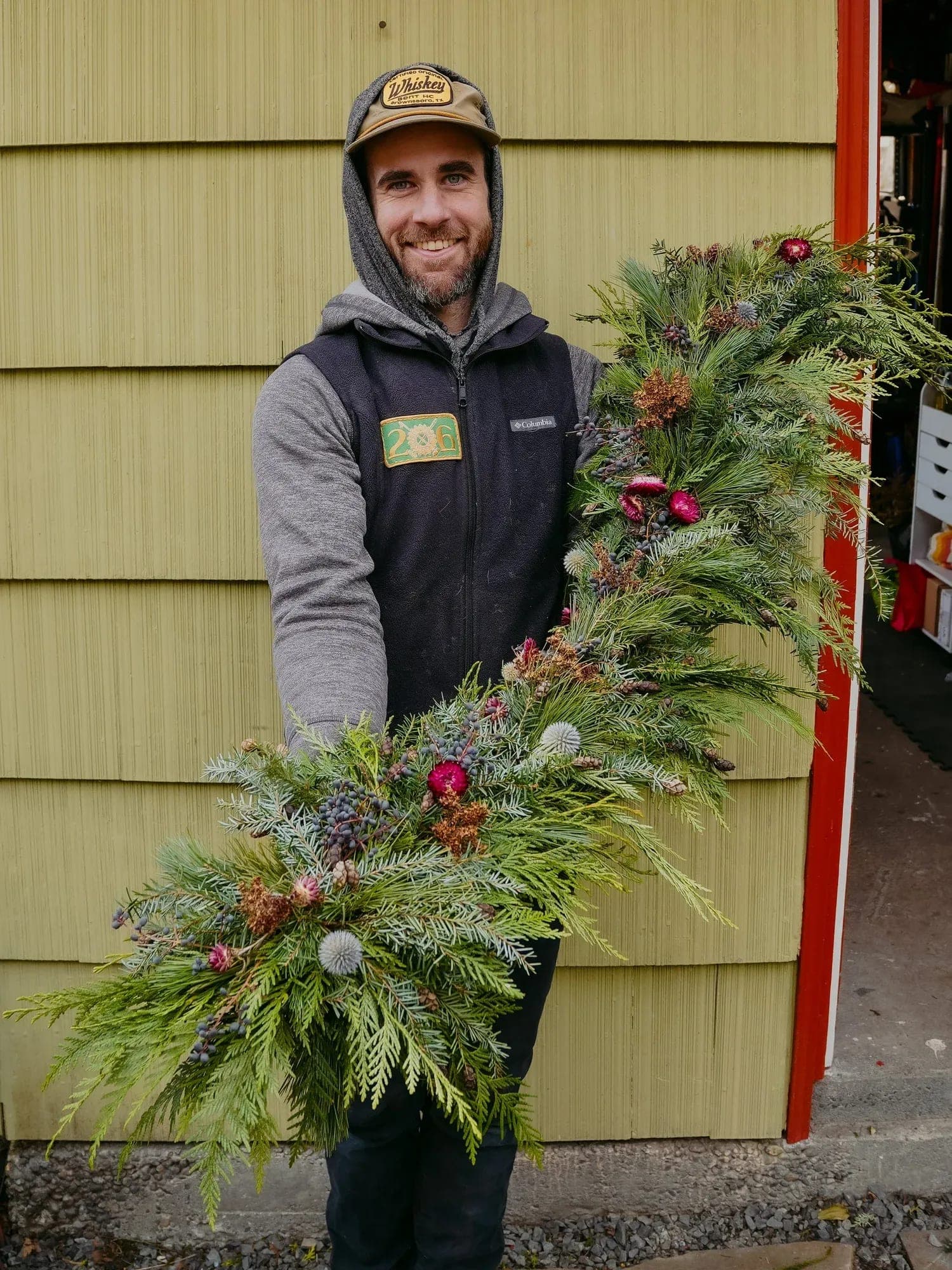 A maker holding a long evergreen table runner draped across both arms, dressed with cedar, thistle, cones and burgundy berries.