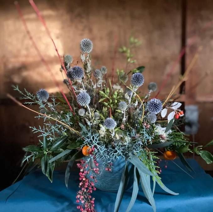 A winter table arrangement of blue globe thistle, silver cedar, eucalyptus and trailing pink peppercorn in a stone vase, on a teal cloth.