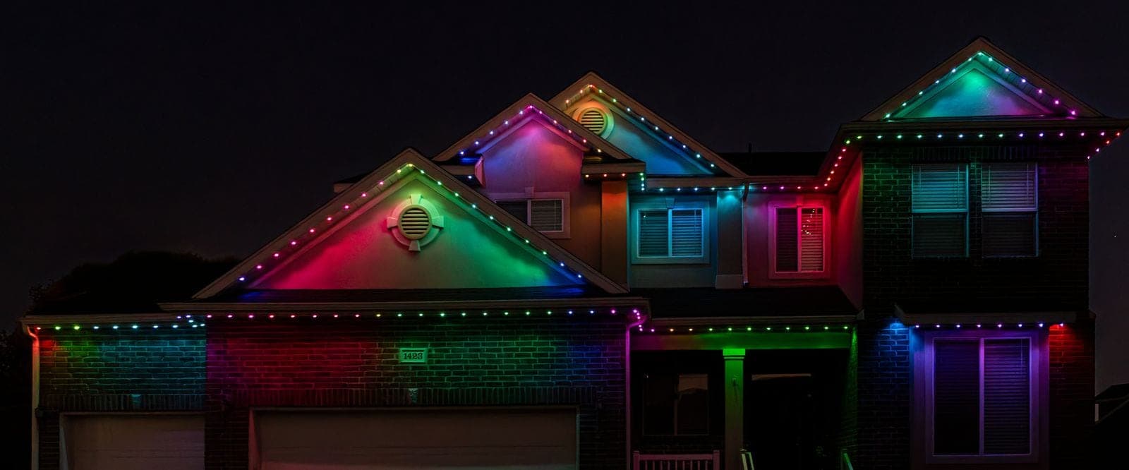 A two-story home at night with programmable roofline lighting running every gable and eave in magenta, green, teal and blue, the brick and stucco washed in the same colors.