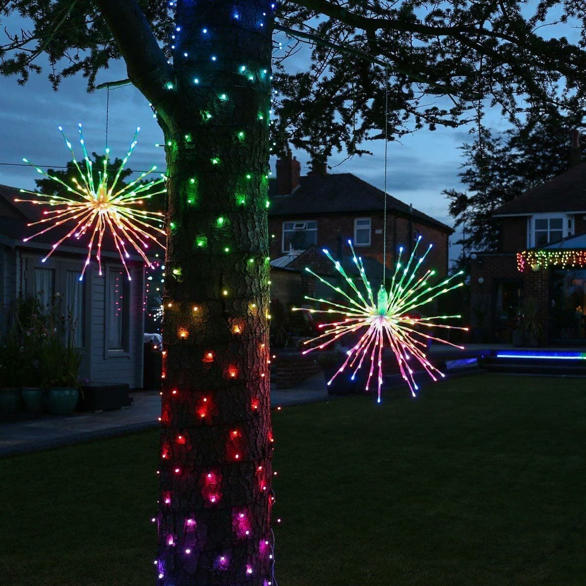 Multicolor spritzer starbursts mounted up a tree trunk at night with a lit yard behind.