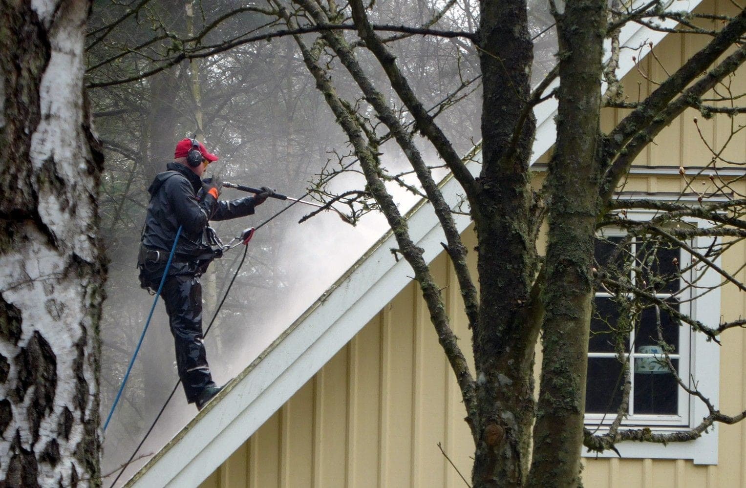 A worker in rain gear blasting a steep roof with a pressure washer wand, throwing up a cloud of spray