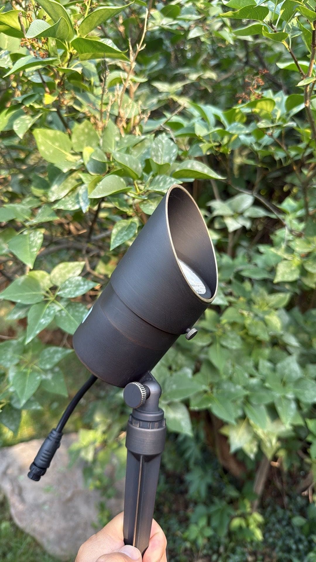 A bronze bullet spotlight on a ground stake with its connector lead, held in front of garden foliage.