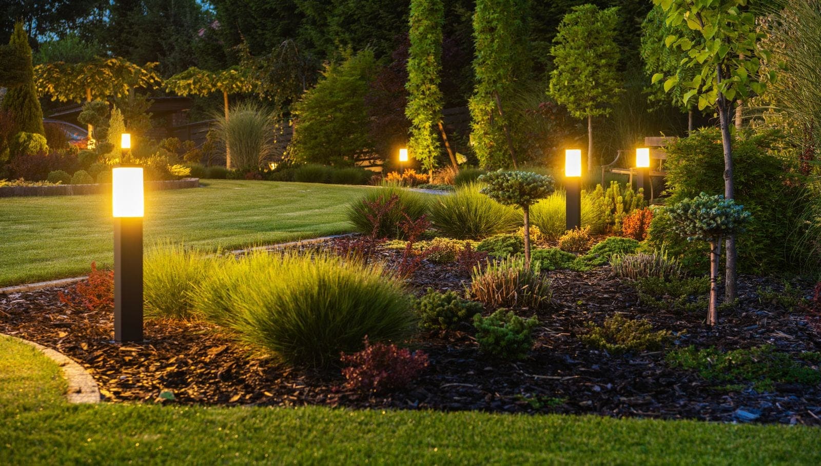 Square bollard lights spaced along the edge of a lawn at dusk, throwing warm pools over bark beds, ornamental grasses and clipped shrubs.