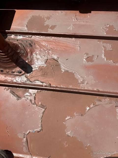 Red paint peeling and flaking off a metal roof panel in large patches, exposing the substrate beneath.