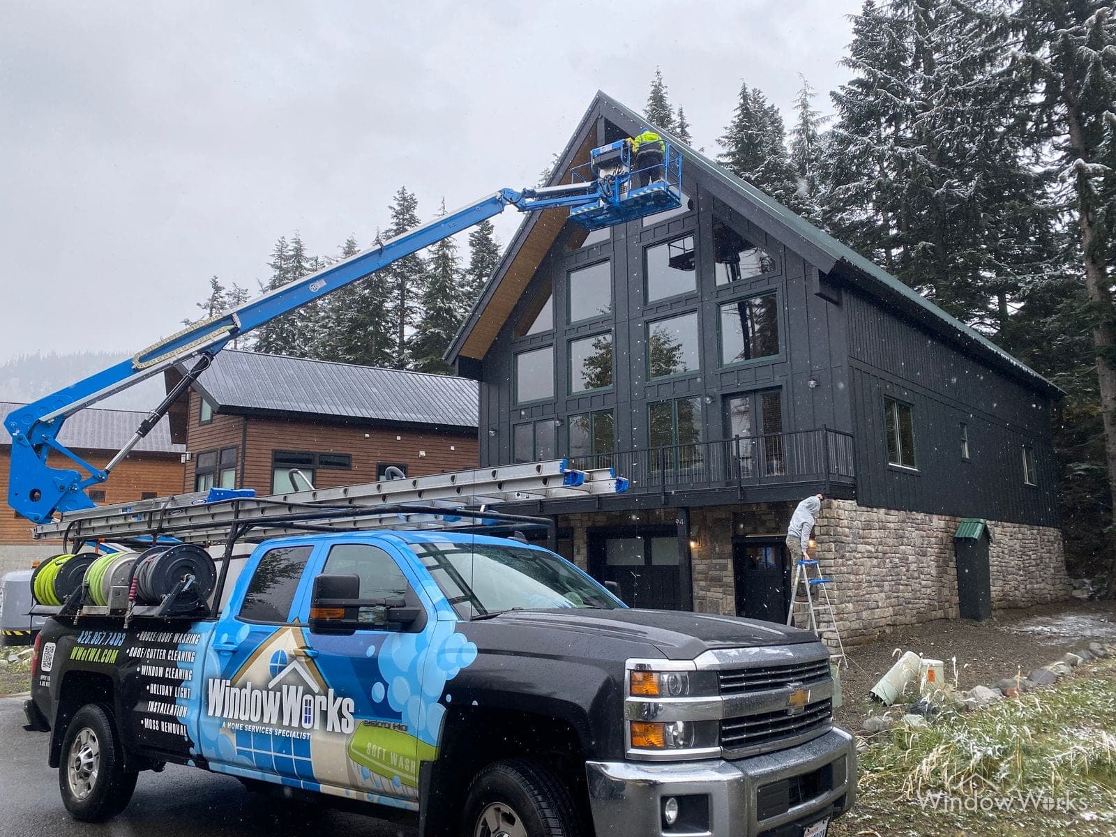 A WindowWorks technician working from a boom lift at the peak of a dark A-frame mountain home during snowfall, the service truck parked below