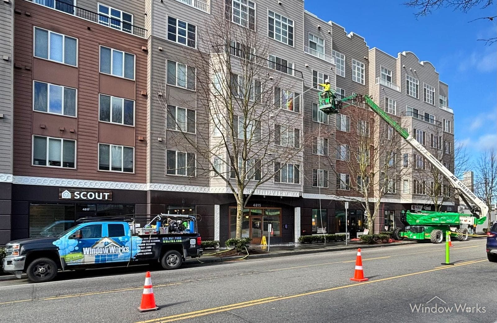 A boom lift raised to the top floor of a five-storey apartment block on a clear day, the WindowWorks truck and traffic cones set out along the closed lane below