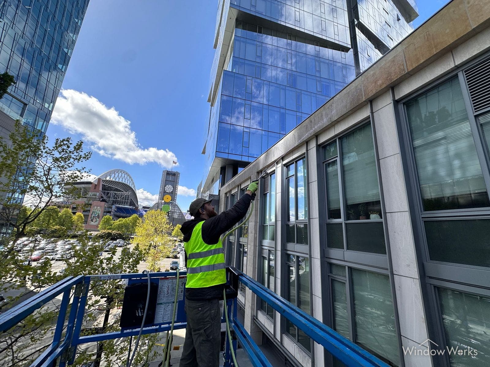 A WindowWorks technician in a high-visibility vest working from a boom lift basket, washing the stone and glass facade of a downtown Seattle office building with Lumen Field behind him