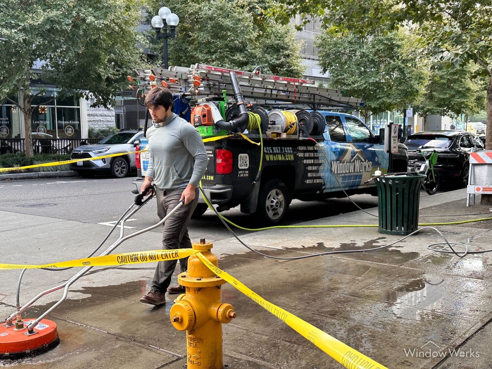 A WindowWorks technician running a surface cleaner along a downtown Seattle sidewalk beside the branded service truck, the work area taped off and the cleaned concrete running out behind him