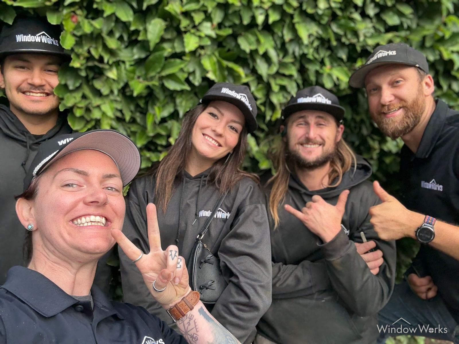 Five WindowWorks team members in company caps, hoodies and polos grinning in front of an ivy covered wall, throwing a peace sign, a shaka and a thumbs up