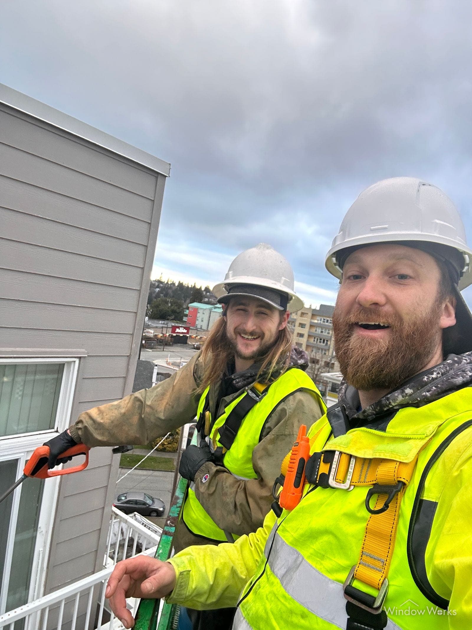 Two WindowWorks technicians in hard hats and high-visibility vests grinning from a lift beside the building siding they are washing