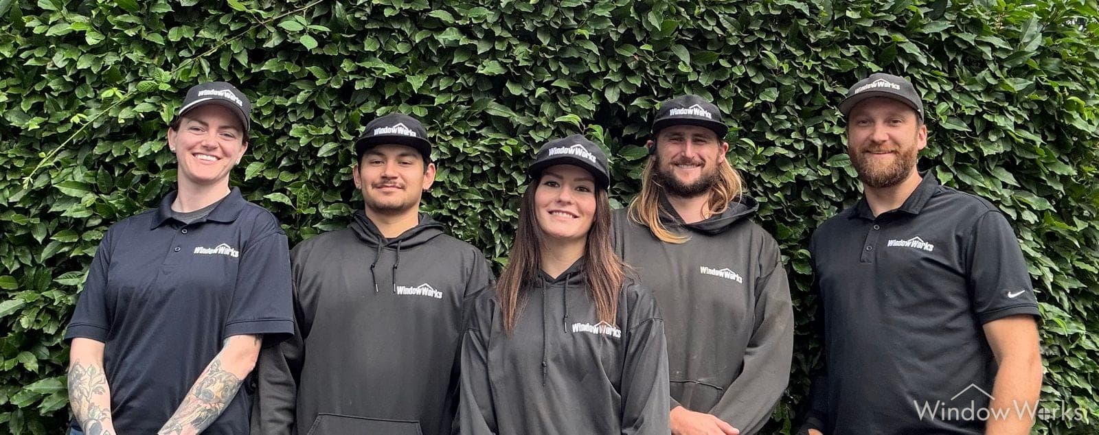 Five WindowWorks technicians in company caps and shirts standing shoulder to shoulder in front of an ivy wall, the crew a customer meets on service day.