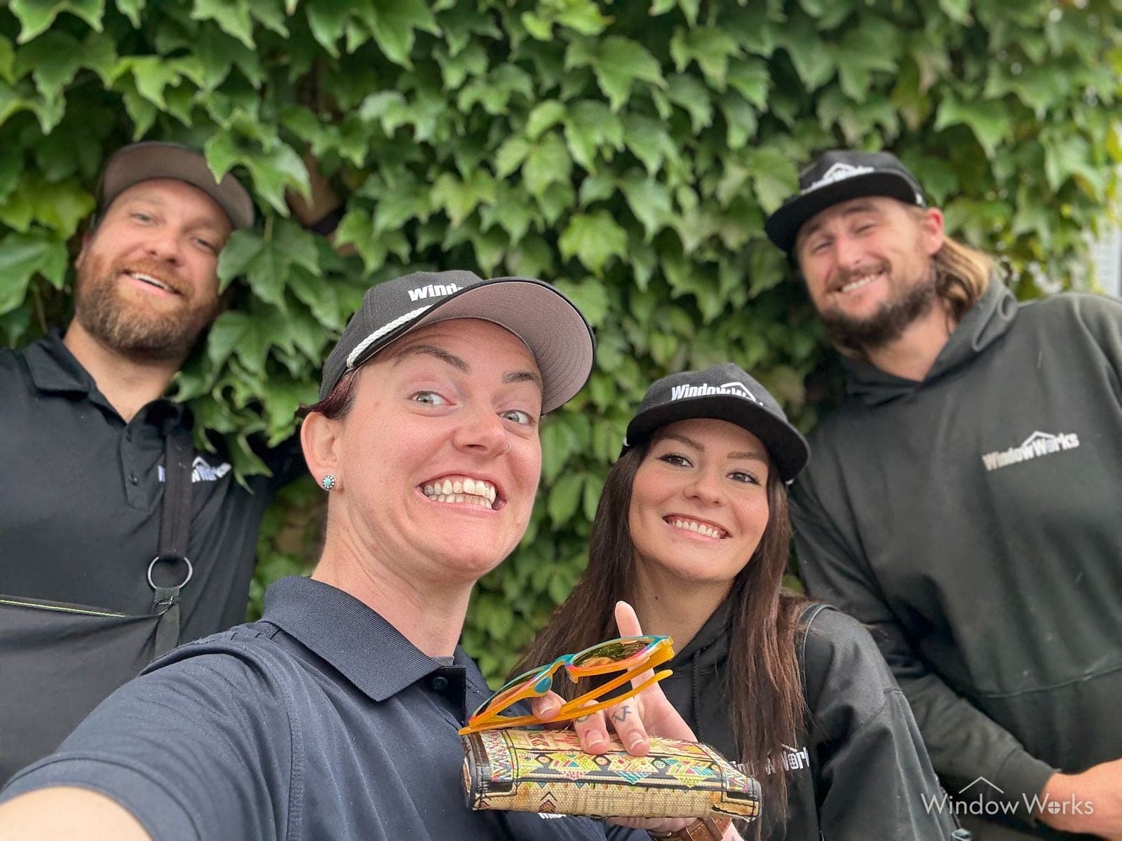 Four WindowWorks team members in company caps, polos and hoodies grinning for a selfie in front of an ivy covered wall