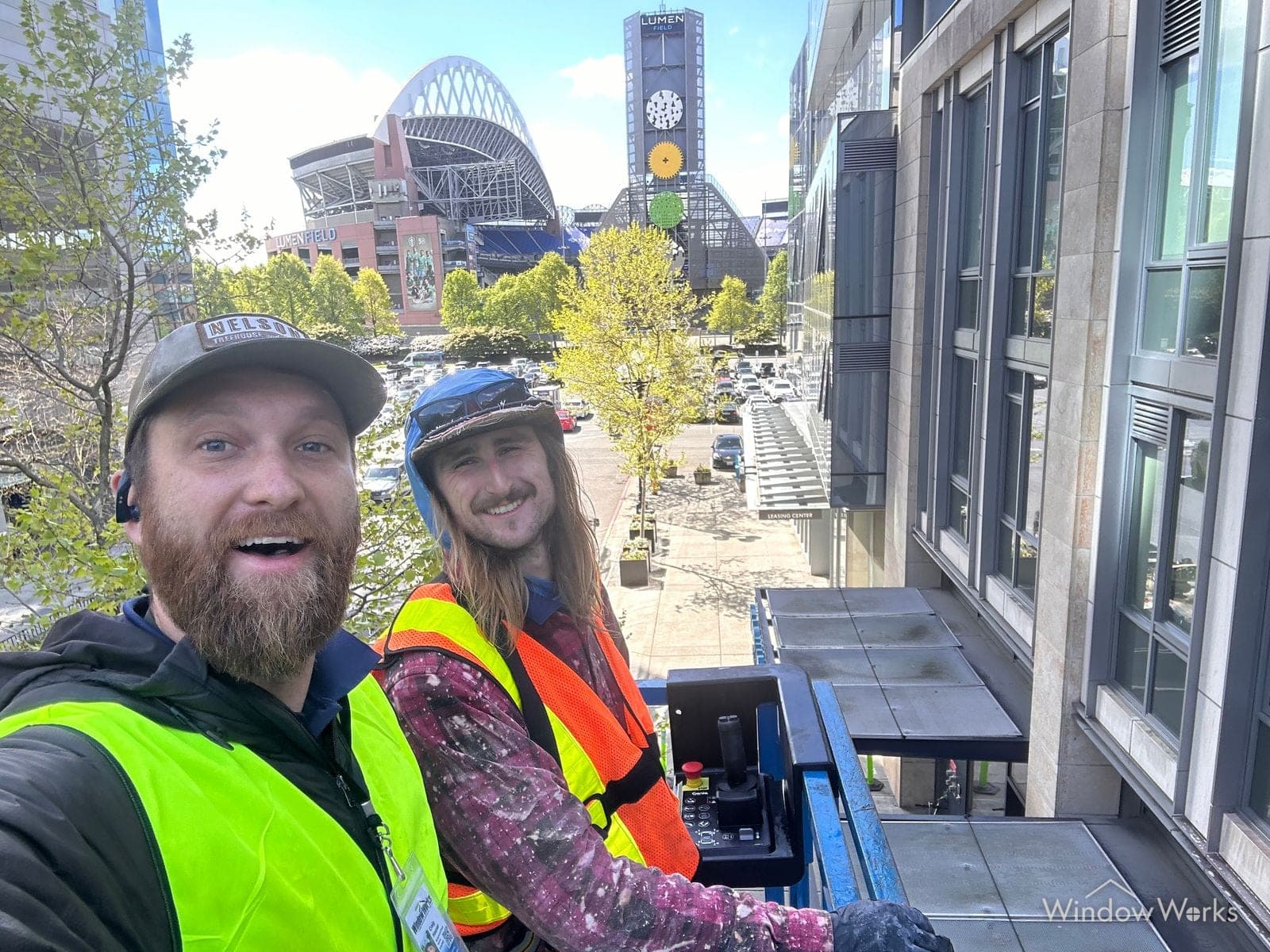 Two WindowWorks technicians in high-visibility vests grinning from a lift in downtown Seattle, the stadiums behind them