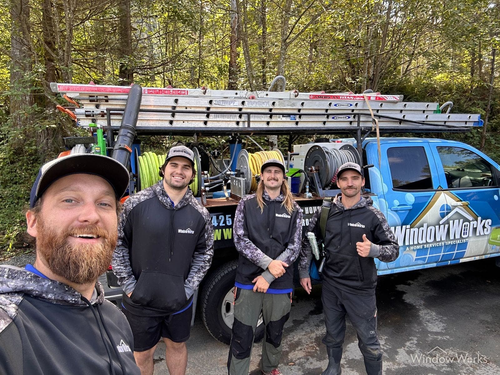 Four WindowWorks technicians gathered at the loaded service truck at a wooded Eastside jobsite, ladders and hose reels racked behind them
