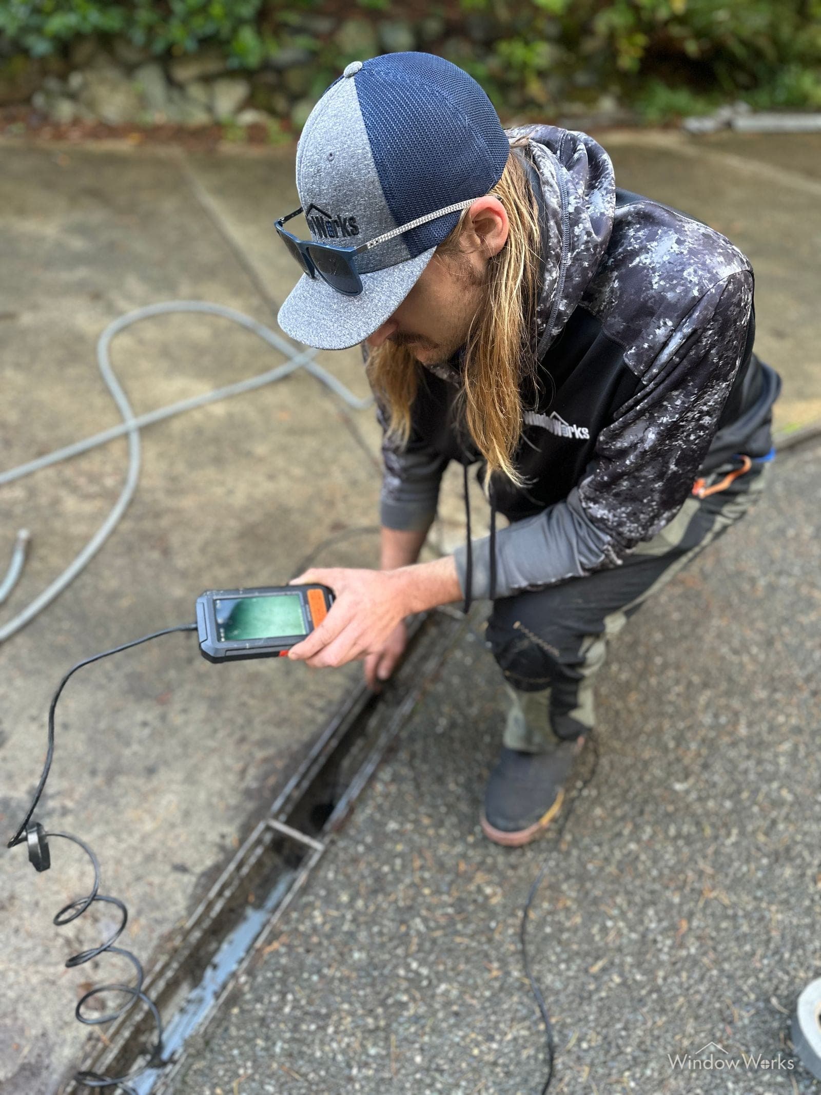 A WindowWorks technician crouched at a driveway trench drain, watching the inspection camera monitor as the line is fed into the buried pipe on the Eastside