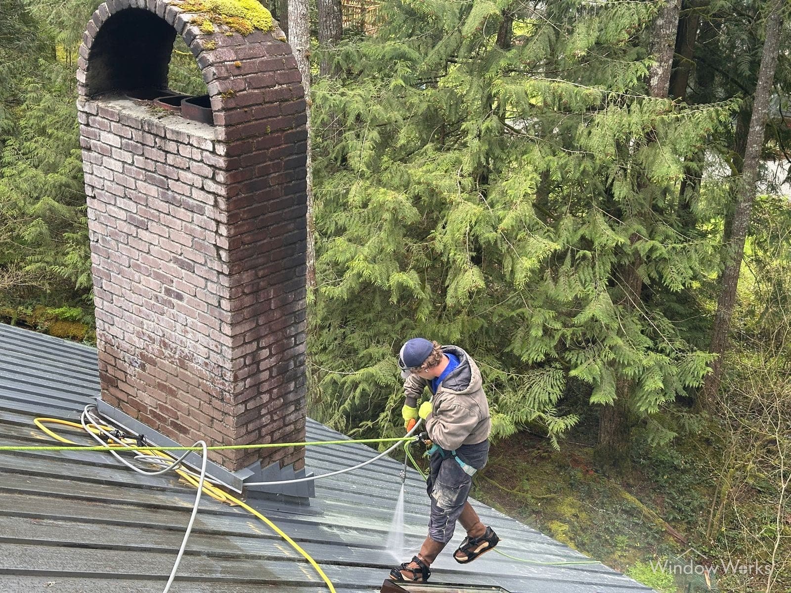 A WindowWorks technician soft washing a standing-seam metal roof beside a tall brick chimney at a home surrounded by evergreens